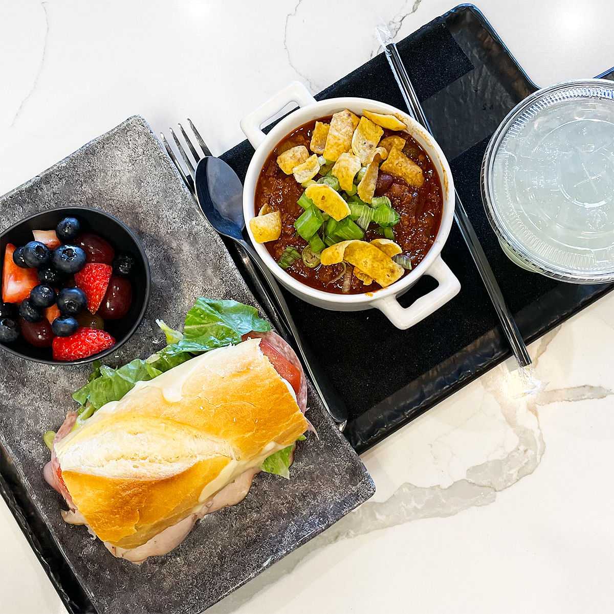 Sandwich, berries, and a bowl of chili with garnish served on a tray, accompanied by a cup of beverage.